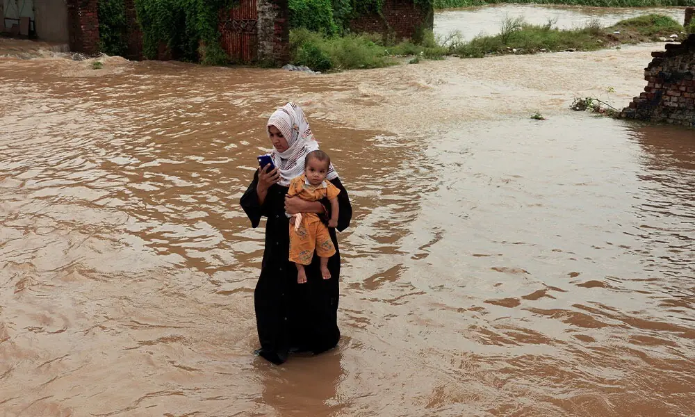  A woman uses a mobile phone as she carries her baby along a flooded street, following monsoon rains and rising water levels in Sialkot, Punjab province, Pakistan, August 27, 2025. REUTERS/Akhtar Soomro 